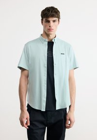 Νέος άνδρας που φοράει ανοιχτό μπλε πουκάμισο με κοντό μανίκι πάνω από μαύρο T-shirt με το λογότυπο "BOSS", σε συνδυασμό με μαύρο παντελόνι, στέκεται μπροστά σε λευκό φόντο.