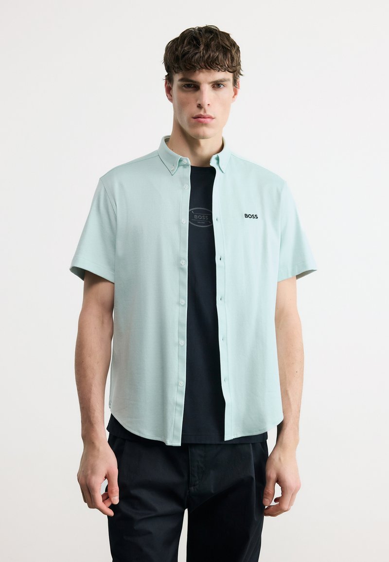 Νέος άνδρας που φοράει ανοιχτό μπλε πουκάμισο με κοντό μανίκι πάνω από μαύρο T-shirt με το λογότυπο "BOSS", σε συνδυασμό με μαύρο παντελόνι, στέκεται μπροστά σε λευκό φόντο.