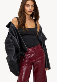 Femme aux longs cheveux bruns portant une veste en cuir noire épaules dénudées, un débardeur noir et un pantalon bordeaux texturé brillant, posant avec assurance.