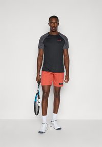 Schwarz-graues Sport-T-Shirt mit rotem Logo, kombiniert mit korallenfarbenen Sportshorts. Das Modell hält einen Tennisschläger und trägt weiße Sportschuhe.