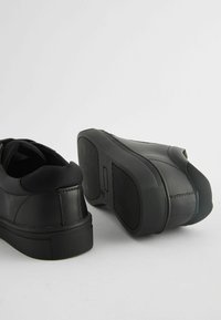 Des baskets noires avec une tige en cuir lisse, un col en textile, un bout arrondi et une semelle en caoutchouc texturée avec un motif en zigzag.