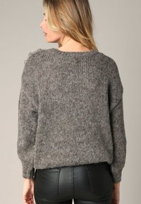 Pull en tricot gris avec une coupe ample, un tissu texturé et des accents de boutons sur l'épaule. Porté sur un pantalon noir ajusté.