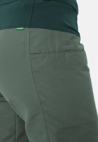 Gros plan sur un pantalon vert foncé avec une poche latérale et une petite étiquette verte portant la mention "Greenshape" près de la ceinture.