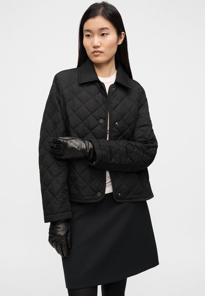 Μαύρο καπιτονέ μπουφάν με κουμπιά, γιακά άνοιγμα και υφή υφάσματος. Φορεμένο πάνω από ένα ανοιχτόχρωμο πουλόβερ με μαύρη φούστα. Μαύρα δερμάτινα γάντια.