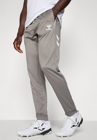 Grijze sportbroek met een gladde textuur, voorzien van witte logo- en chevrondetails aan de zijkant, gecombineerd met witte en zwarte sportschoenen.