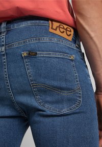 Jeans de denim azul con un parche de cuero de la marca Lee, costuras amarillas en contraste y un clásico diseño de cinco bolsillos.
