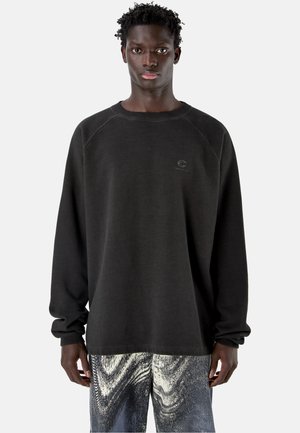 Young man with dark skin wearing a plain black long-sleeve shirt and patterned black-and-white pants standing against a light background.