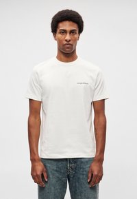 Krátky rukáv krémové bavlnené tričko s malým čiernym vyšívaným logom na ľavej strane hrudníka, v kombinácii so svetlomodrými vyblednutými džínsami.