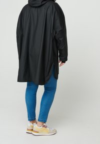 Sort, oversized regnjakke med hætte, der har en rund nederste kant. Samlet med blå jeans og sneakers med gule detaljer. Glat tekstur over hele jakkens overflade.