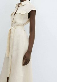 Femme portant une robe beige sans manches à boutons avec des poches poitrine et une ceinture nouée à la taille, posant devant un fond clair uni.