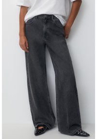 Tummanharmaat, korkeavyötäröiset, leveälahkeiset farkut, joissa haalistunut pinta, klassinen viisitaskumalli ja napillinen etulista.