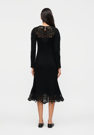 Γυναίκα με μακριά σκούρα μαλλιά που φοράει μαύρο μίντι φόρεμα με δαντελένιες λεπτομέρειες στο επάνω μέρος της πλάτης, στα μανίκια και στο τελείωμα, στέκεται με την πλάτη προς την κάμερα.