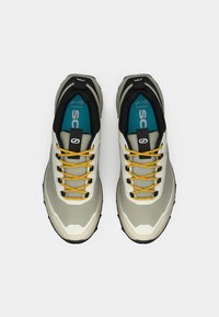 Zapatillas deportivas de cordones con un upper en gris claro y crema, cordones amarillos, detalles en negro y suelas texturizadas. Cuenta con un cuello acolchado para mayor comodidad.