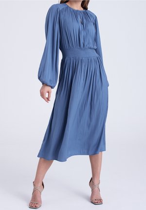 Donna che indossa un vestito midi blu a maniche lunghe con vita stretta e gonna plissettata, abbinato a sandali beige con cinturini e tacco alto.
