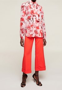 Kvinna som bär en långärmad blus med rosa och röda blommor och en rosett, ljusröda korta breda byxor och svarta högklackade sandaler.