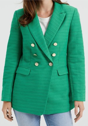 Kvinde iført tekstureret grøn dobbeltradet blazer med seks guldknapper over hvid top og lyseblå jeans, vist fra hals til hofter.