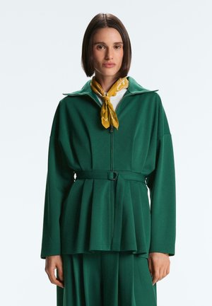 Kobieta o krótkich ciemnych włosach, ubrana w zieloną kurtkę na suwak z paskiem, dopasowaną spódnicę, biały top oraz żółty szal z wzorem, zawiązany wokół szyi.