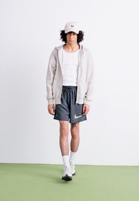 Lys grå zip-up hættetrøje, hvid tanktop, sorte shorts med grønt Nike-logo, hvide sneakers og beige cap. Krøllet hår synligt.