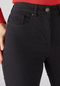 Jeans de mezclilla negra con una textura suave, cintura alta y diseño de cinco bolsillos. Presentan un cierre frontal con botón y detalles de costura sutil.