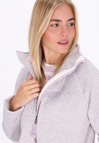 Donna bionda che indossa una giacca in pile di un leggero viola con foderatura in sherpa bianco, parzialmente zipata, mentre guarda di lato.