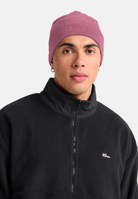 Jonge man met een mauve fleece beanie en een zwarte Jack Wolfskin fleecejack met rits, met ringoorbellen en een neutrale uitdrukking.