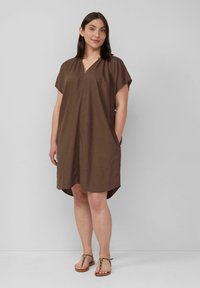 Bruine, korte jurk van lichtgewicht stof met een V-hals en zijzakken. Asymmetrische zoom en een relaxte pasvorm. G gedragen met platte, gespen sandalen.