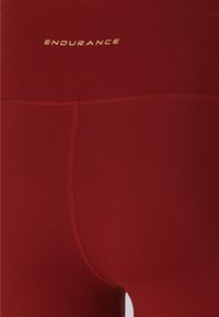 Rode sportlegging met een gladde textuur, voorzien van een hoge tailleband en een gouden "ENDURANCE" logo. Zichtbare naden benadrukken het ontwerp.