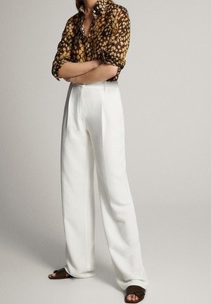 Person, die mit verschränkten Armen ein braun gemustertes Hemd, weiße, weit geschnittene Hosen und braune Sandalen mit offenem Zehenbereich vor einem einfarbigen Hintergrund trägt.