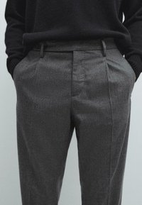 Pantaloni in misto lana grigi con vestibilità tailored, caratterizzati da pince anteriori, due tasche laterali e una trama liscia. Indossati con un maglione nero.