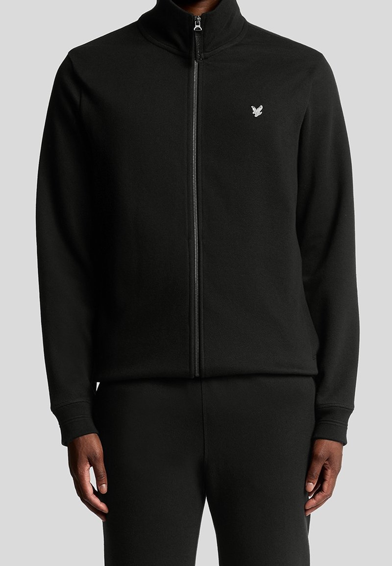 Homme portant une veste noire zippée avec un petit logo d'oiseau blanc sur le côté gauche de la poitrine, assortie à un pantalon noir.