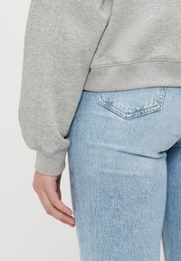 Szürke kapucnis pulóver bordás mandzsettákkal, világoskék, enyhén koptatott skinny farmernadrággal párosítva. A farmernadrágon egy kis hátsó zsebdísz található.