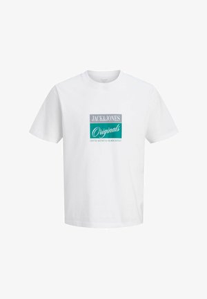 Adolescente con capelli ricci che indossa una t-shirt bianca con grafica, jeans blu a taglio largo e scarpe marroni, in piedi su sfondo bianco.
