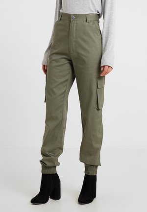 Kvinde iført olivengrønne cargobukser med elastiske manchetter og sorte ankelstøvler med hæl, kombineret med en lysegrå langærmet bluse.