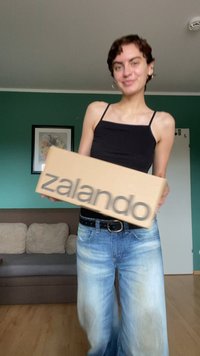 Caja de cartón con el logo de Zalando, sostenida por una persona que lleva una camiseta sin mangas negra y unos pantalones vaqueros de pierna ancha azul claro, en una habitación con paredes verdes.