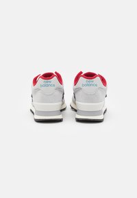 Pár šedobílých tenisek New Balance s červeným vnitřním podšíváním, viděný zezadu na bílém pozadí.