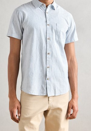 Man wearing light blue short-sleeve button-up shirt with thin vertical stripes and beige pants, standing against neutral background.