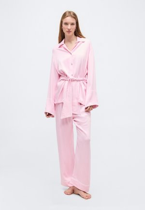 Γυναίκα με μακριά μαλλιά που φοράει άνετο ανοιχτό ροζ σατέν πουκάμισο και αντίστοιχο παντελόνι με φαρδιά μπατζάκια, όρθια ξυπόλητη σε ένα απλό ανοιχτό φόντο.