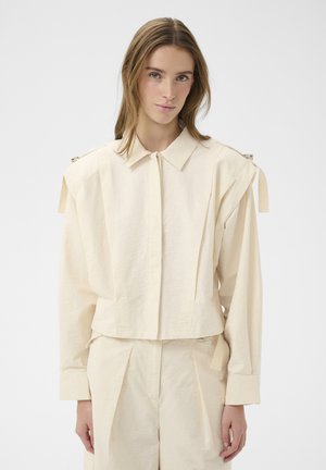 Frau trägt ein lockeres, cremeweißes Langarmshirt mit Schulterriemen und passende hochtaillierte Hose vor einfachem Hintergrund.