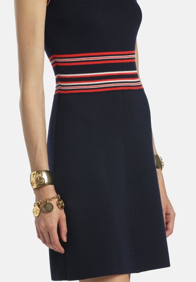 Námornícke rebrované pletené šaty s červenými a bielymi horizontálnymi pruhmi v páse; doplnené zlatým náramkom a náramkom s príveskami.
