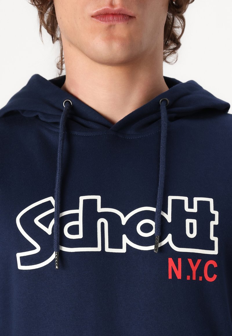 Gros plan sur une personne portant un hoodie bleu marine avec le texte "Schott" en blanc surligné et "N.Y.C" en rouge imprimé sur le devant.