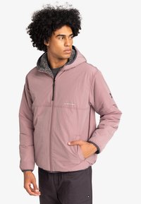 Veste légère zippée en rose pâle avec une finition texturée, dotée d'une capuche et d'une doublure en polaire grise. Comprend des poches latérales et des poignets élastiques.