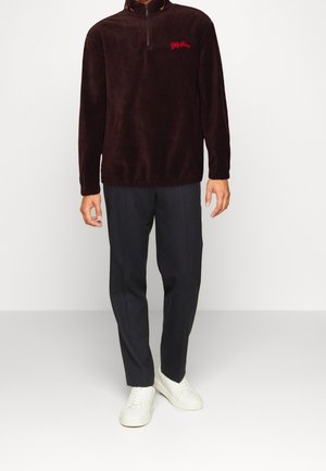 Mand iført en mørkebrun fleece-pullover med lynlås og rød logo, sorte bukser og hvide sneakers mod en ensartet lys baggrund.