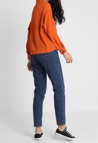 Orange langærmet bluse, løs pasform med knaplukning ved håndleddene, parret med mørkeblå lige ben jeans og sorte kanvas sneakers.