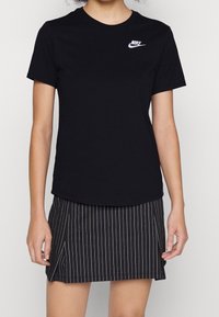 Kvinna iklädd en svart Nike-t-shirt med vitt logotyp och en svart randig kjol med sprund framtill, stående mot en enkel bakgrund.