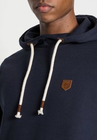 Navy modrá mikina z měkké látky, s klokankou, krémovými šňůrkami s hnědými koženými detaily a koženým logem.