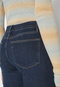 Vaqueros de mezclilla azul oscuro con dos bolsillos traseros, costuras visibles y una forma ajustada. Acompañados de un suéter de tejido ribeteado multicolor.