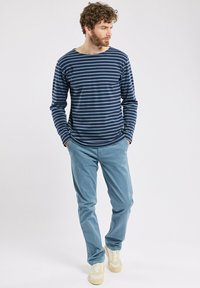 Camicia a maniche lunghe a righe blu navy e azzurro abbinata a pantaloni azzurri e sneakers beige, in piedi su uno sfondo neutro.
