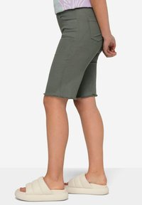 Shorts de mezclilla verde oliva con dobladillo deshilachado, ajuste slim, bolsillos laterales y una textura suave, combinados con sandalias blancas sin talón.
