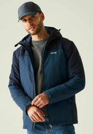 Hombre con chaqueta con capucha azul marino, suéter gris, vaqueros azules y gorra de béisbol azul marino, ajustándose los puños de la chaqueta.