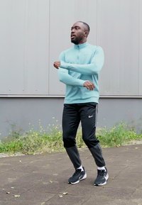 Sportstøj bestående af en lyseblå langærmet top og sorte bukser med grå detaljer, samt sorte sportssko på en belagt overflade.